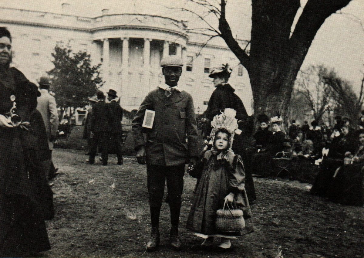 Black and white photo of a man in a suit jacket and pants with a young girl in a dress and hat, in front of the White House.