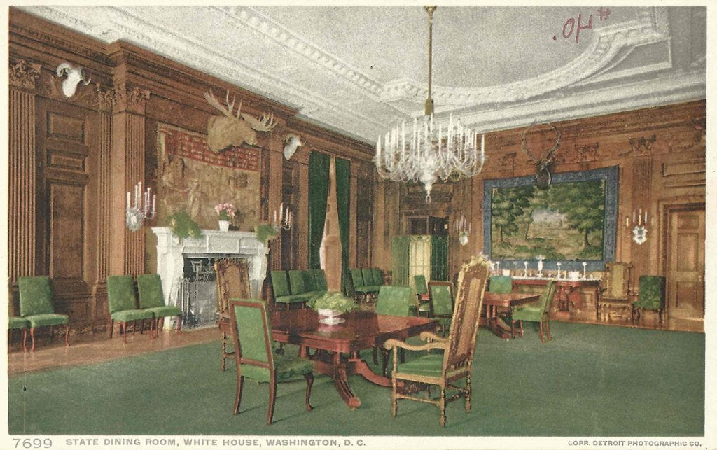 Postcard of the State Dining Room. It has dark wood walls with green carpeting and wooden furniture with green upholstery. 