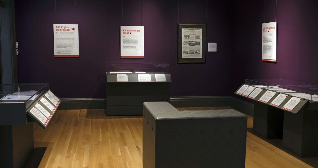 photograph of a room with dark purple walls and wood floors. In the room are display cases holding objects 